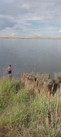 A man in black shorts stands in shallow water near a dirt bank. In the distance, two birds are visible on the water. The sky is cloudy.