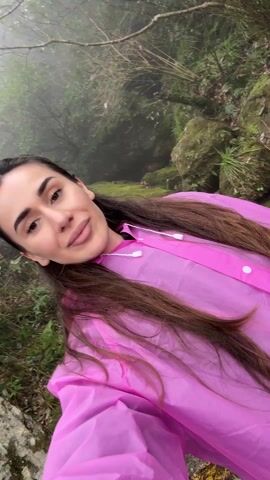 A woman wearing a pink plastic poncho is recording a video of herself in a lush, rocky forest. She is smiling. The background consists of moss-covered rocks and greenery. The day appears overcast and possibly misty.