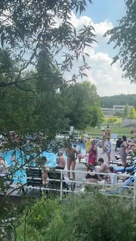 The video shows a daytime scene at an outdoor swimming pool area. Several people are visible, some in swimsuits, lounging on chairs or standing near the pool. The pool itself is filled with blue water, and there are people swimming in it. Surrounding the pool is a paved area with lounge chairs and some greenery, including trees and bushes. In the background, beyond the pool area, there is a body of water, possibly a lake or pond, surrounded by dense trees. The sky is visible with scattered clouds. The camera pans slightly, offering different views of the pool and the surrounding environment.