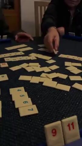 People are playing the game Rummikub. The table is covered with scattered tiles and numbered tile holders with tiles arranged in them.  A person on the right is moving tiles around. A person on the left is holding a cell phone.  The scene is indoors with a wooden cabinet in the background.