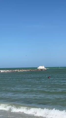 The scene is a view of a calm sea on a bright, sunny day. A long breakwater made of rocks stretches across the middle ground of the frame. On the far side of the breakwater, a white boat is docked. In the foreground, small waves break on the beach, and a person is swimming in the water. A small dark bird flies high in the clear blue sky.