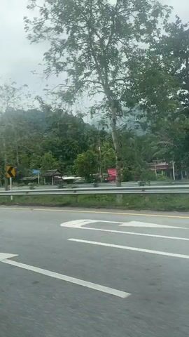 The video shows a view from a car traveling on a highway on a cloudy day.  The highway has white lane markings and a yellow line on the shoulder.  On the side of the road, there is a guardrail and a grassy area, followed by trees and vegetation.  In the background, there are mountains covered in lush green foliage.  Road signs are visible, including a yellow and black chevron sign indicating a curve and a blue sign with a white arrow pointing to the left, indicating a U-turn. Several cars are seen on a cross street to the right. A roundabout is seen with alternating black and white paint.  Streetlights are present along the roadside.