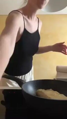 A fair-skinned woman stands in a kitchen, cooking something in a frying pan. She wears a black tank top and light-colored pants. She uses a spatula to flip the food in the pan. The kitchen has yellow walls and a white ceiling light. The pan is black and sits on a black stovetop. A metal stepladder and a beige couch are visible in the background.