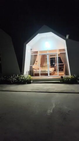 The video shows a nighttime view of a row of small, modern, white structures with triangular roofs. The structures are illuminated from within and without, casting a warm glow into the dark surroundings. In front of the structures, there is a paved area with a few parked vehicles, including a white car and a motorcycle. Some small, decorative plants line the walkway in front of the buildings. One of the structures has large glass doors and windows, revealing a glimpse of a bedroom inside, with a bed and curtains. The overall impression is one of a modern, possibly boutique, lodging or accommodation facility.