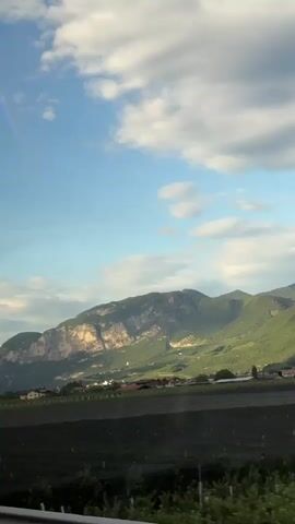 A landscape video captures rolling green hills and mountains under a partly cloudy sky. In the foreground, there are fields of green crops and a hint of a fence or railing. Further back, several houses with red roofs are visible, nestled among the trees and on the lower slopes of the mountains. The sunlight casts shadows across the terrain, highlighting the contours of the landscape. The overall impression is one of a rural, natural setting.