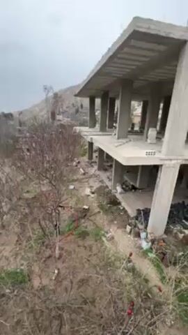The video shows an unfinished concrete structure standing in a rural area. The building is two stories tall and appears to be a residence under construction. The unfinished state reveals the concrete beams and pillars that support the floors and roof. The surrounding environment is characterized by sparse vegetation, including trees without leaves, possibly indicating a winter or early spring setting. A hill or small mountain is visible in the background. Debris and construction materials are scattered around the base of the building. The sky overhead is overcast.