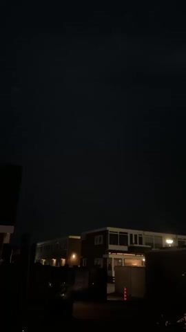 The video captures a dark exterior scene at night, with a low-rise building in the background. The building appears to be illuminated by interior lights, casting a warm glow through its windows. A dark, silhouetted foreground suggests the presence of vegetation or other structures. The overall atmosphere is one of quiet darkness, with the building being the only source of significant light. The sky is a deep black, indicating nighttime. No people or animals are visible in the frame.