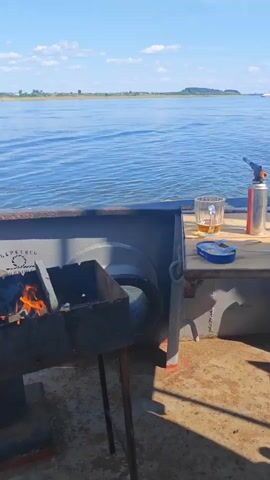 The video shows a daytime scene on a body of water, possibly a lake or river. In the foreground, a small grill with hot coals is visible on the left. A wooden table is set up with two glasses containing amber liquid, a gas torch, a small blue object that appears to be a lighter, and a small metal utensil. To the right of the table, a can is partially visible. The camera pans across the water, revealing a distant shoreline with vegetation and some structures. The sky is blue with scattered white clouds. The water is calm with small ripples. There is a gentle movement of the boat or platform from which the video is filmed. No people are visible in the frame.