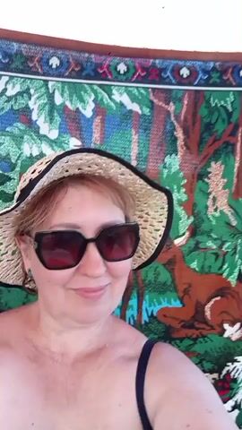 A woman wearing a straw hat and sunglasses lies on her back on a colorful tapestry. The tapestry depicts a forest scene with trees and a deer. The woman is smiling and looking at the camera. The video captures the woman from the chest up.