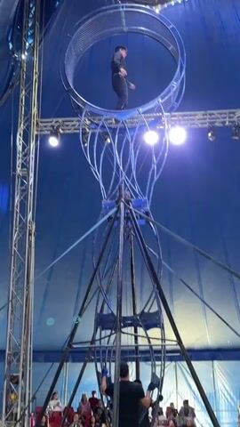 A male circus performer stands at the top of a tall, elaborate metallic structure that culminates in a large spinning wheel. He is dressed in a black outfit with decorative trim on the collar and chest. The performer gestures with his arms as he balances in the wheel, which is suspended high above the ground within a circus tent. The tent's blue fabric is visible in the background, along with stage lighting. The performer appears to be in the middle of a routine, with the wheel rotating around him.