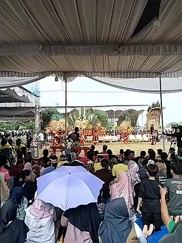 A performance is underway beneath a large white tent. The stage is decorated with what appear to be large, stylized depictions of trees and possibly a lion. Several performers are on the stage, some dressed in colorful costumes. In the foreground, an audience of people sits on the ground, facing the stage. Many of the audience members are wearing head coverings, suggesting a cultural or religious context. One person in the audience is holding a purple umbrella. The overall atmosphere appears to be one of a community gathering or festival.