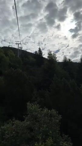 The video opens looking up at a cloudy sky with the sun peeking through, over a dense forest of dark green coniferous trees on a mountainside. A single ski lift tower is visible on the left side of the frame, with a cable extending out of view. The camera then pans to the right, revealing a wider view of the mountainous landscape. Rolling green hills stretch into the distance, dotted with small houses and a winding road. The sky remains heavily clouded. The camera continues to pan right, showing more of the green hills and the houses nestled within them. The video concludes by panning back to the left, returning to the view of the forested mountainside and the cloudy sky.