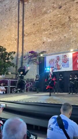 This video captures a vibrant folk dance performance on an outdoor stage. The central subjects are a group of dancers, some in black, traditional-looking costumes with red accents, and others in black outfits with fur hats. One dancer in the foreground, wearing a red headscarf and black and red attire, is mid-twirl, her skirt fanning out dramatically. Another dancer in a black costume with a tall, dark fur hat is also actively performing, his movement suggesting a dynamic dance. Several other dancers in similar black outfits are positioned behind them, some holding what appear to be props, possibly swords, adding to the intensity of the performance.

The setting is an outdoor stage at dusk or early evening, indicated by the artificial lighting on stage and the general ambient light. Behind the dancers, a large banner displays text. The prominent text reads "Calella Folk Festival" along with smaller text and logos, including social media icons for Facebook and Instagram, and the location "Ajuntament de Calella." Images of performers in traditional costumes are also visible on the banner. The backdrop of the stage is a textured, light-colored stone wall, likely part of a historic building. Purple bougainvillea cascades down from above, adding a touch of natural beauty to the scene. In the foreground, the backs of audience members are visible, one wearing a white t-shirt with the text "folkt" and "festival" visible, suggesting they are attending the event. The overall mood is lively and celebratory, with the dancers exhibiting energy and skill.