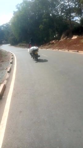 A motorcycle is parked on the side of a paved road. The road curves around a bend.  A forest is on the left side of the road. The sky is visible. The camera appears to be angled slightly. A concrete barrier lines the road. The ground beside the road is brown.