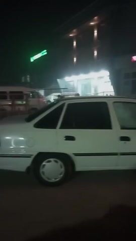 At night, a white car, possibly a Daewoo, is parked on the street. The car has black trim. Behind it is a building that looks like a store or office with a sign that says "akfa" in red letters. There is also another building further away with a green sign. It appears to be foggy or hazy. The lighting is dim. There are no people clearly visible in the video.