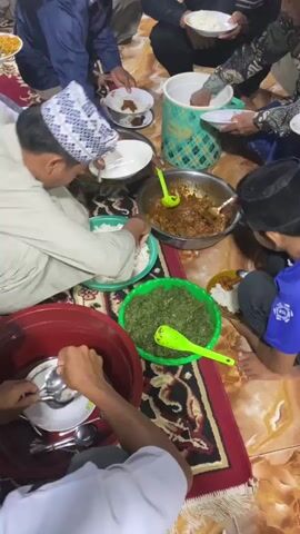 A group of people are gathered on the floor around several bowls of food. The floor is covered with a rug. The people are dressed in traditional attire and appear to be eating together. One person is serving food from a large bowl into a smaller bowl. Another person is taking food from a bowl with a spoon. There are several bowls of food, including rice, noodles, and a stew-like dish. A large blue and green container is in the background. The lighting is dim.