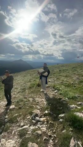 The video shows two men standing on a grassy, rocky hillside. One man is looking at and using his phone, while the other is playing a wind instrument, possibly a bagpipe, with a trekking pole next to him. The sun is shining brightly, partially obscured by clouds. In the background, there are mountains.