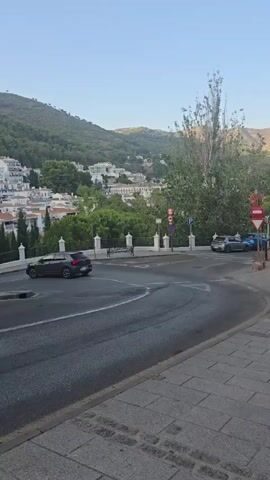The video captures a street scene with a white car driving away from the camera on a curved road. The car is in the foreground, with its rear visible as it turns to the right. The road is paved and appears to have tire marks. In the background, a white town with red-tiled roofs is nestled on a hillside covered in green trees. The sky is clear and blue. There are some trees and white decorative posts along the side of the road. A traffic sign is visible on the right side of the road. Another car, possibly blue, is parked or driving in the distance. The overall scene suggests a sunny day in a Mediterranean town.