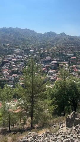 This is a vertical, outdoor, handheld video taken on a sunny day. The camera is positioned on a rocky, elevated viewpoint overlooking a village nestled in a valley between rolling, vegetated hills. In the foreground, there are rocks and several pine trees, with one prominent tall pine tree in the center. The village below consists of numerous houses with red-tiled roofs. The sky is clear and blue. The video pans slightly to the right, focusing on a metal signpost with three plaques. The plaques contain text in a non-English language, likely Greek, with names and dates. The text appears to be a memorial.