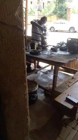 A man wearing a striped shirt and dark shorts works at a cluttered wooden workbench in an outdoor or semi-outdoor workspace. He appears to be using a tool to work on a piece of machinery or an engine part. The workbench is covered with tools and spare parts. Behind the workbench, there is a white car visible in the background. Next to the workbench, there is a black bucket. The video is shot from inside the workspace, with a thick wooden post visible on the left side of the frame. Another person is partially visible on the right-hand side of the frame.