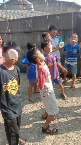 A group of about 12 children, mostly boys, are participating in a game in an outdoor area. Each child has a large, round, white cracker tied to a string, hanging in front of their face. The children are trying to eat the cracker without using their hands, tilting their heads back and opening their mouths wide. Some children are more successful than others. They are all standing on a rough, dusty surface, possibly a courtyard or alleyway. Behind the children is a concrete wall with some markings on it. In the background, there is a metal fence and a glimpse of rooftops and trees. The overall atmosphere seems playful and competitive. The camera captures the scene at eye-level, with slight movements following the action.
