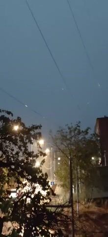 This is a short video clip, filmed at night with a handheld camera, looking up at a dark, overcast sky. Power lines are visible crisscrossing the sky. Below the power lines, there are streetlights illuminating a road, and several trees. The trees in the foreground are silhouetted against the light. There are some faint lights, possibly stars or distant objects, visible in the sky. The overall atmosphere is moody and somewhat atmospheric due to the low light and the presence of the power lines and streetlights.