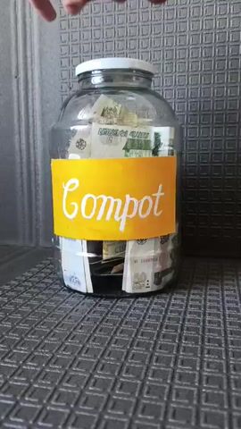 A hand is shown unscrewing the lid of a large glass jar filled with Russian currency, both banknotes and coins. A yellow label with the word "Compot" in white cursive lettering is affixed to the front of the jar. The jar is placed on a textured dark gray surface, possibly a mat, with a light gray patterned wall in the background. The lighting is diffuse and appears to come from the front.