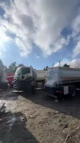 Here is a description of the video:

The video shows a series of tanker trucks in a muddy area. The ground is dark and uneven, suggesting a dirt or gravel surface. Some of the trucks are white with green and blue markings, while others appear older and more worn, one being gray and rusty. A person is seen standing next to one of the white trucks and another person walking towards an older gray truck. Power lines and sparse trees are visible in the background, under a sky with white clouds and a bright sun shining through. Overall, the scene looks like a staging or parking area for these utility vehicles.