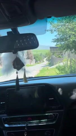The video shows a view from inside a car parked on a narrow street. The street is lined with houses and fences, and green fields can be seen in the distance. A mountain can be seen in the far distance. There is a fluffy white car ornament hanging from the rearview mirror, and beads are resting on the dashboard. The car's infotainment system is visible but not activated. The sky is clear and blue, suggesting it is a sunny day.