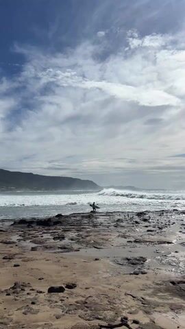 The video shows a beach with a surfer carrying a board standing on a rocky outcrop in the foreground. In the background, large waves are crashing onto the shore, and a green, mountainous landmass stretches across the horizon. The sky above is partly cloudy with patches of blue. The beach appears to be a mix of sand and exposed rock, with small pools of water scattered around. The lone surfer is the only visible person besides those further out in the water. The overall impression is one of a moderately windy day at the beach, with a focus on the surfer's potential interaction with the waves.