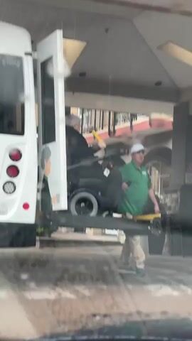 A white bus with a wheelchair lift is parked under a covered entryway of a building on a rainy day. An elderly individual seated in a black power wheelchair with glasses and a dark-colored coat is being lowered on the lift. A man in a green polo shirt, khaki pants, and a white baseball cap is standing beside the lift, holding a remote control and looking toward the person in the wheelchair.  The background shows parts of the building facade and a "No Parking" sign.