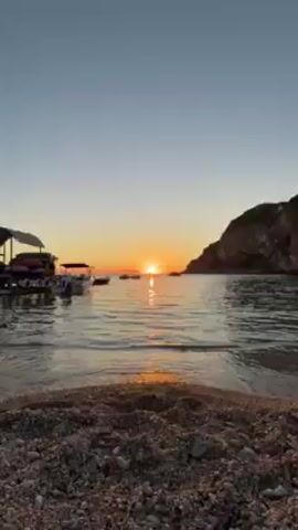 The video shows a beach with a calm sea at sunset. The sun is setting in the middle of the frame, behind a small island. The sky is orange, and the sun's reflection can be seen in the water. There are several small boats in the water, including one motorboat moored very close to the camera. A rocky outcrop is visible in the background on the right. The waves are gentle.