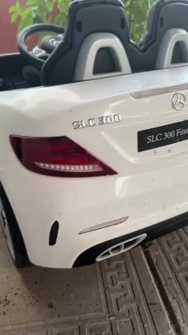 The video shows the rear of a white Mercedes-Benz SLC 300 Final Edition toy car. The car is positioned outdoors on a paved surface, possibly a driveway or patio, with a green leafy plant in the background. The setting appears to be a backyard or garden area, with a glimpse of a fence and possibly a house in the distance.

The car is the central subject, with its taillights, Mercedes-Benz emblem, and "SLC 300" and "SLC 300 Final Edition" badging clearly visible. The car appears a bit dusty, suggesting it has been used outdoors.

There is no activity or event taking place in the video; it is a still shot of the toy car. The time of day is not explicitly indicated, but the lighting suggests it is daytime, likely with moderate weather, as there are no signs of rain or harsh sunlight. The location cues suggest a residential area in rural or suburban Russia. The overall tone is neutral, focusing on the detail of the toy car.