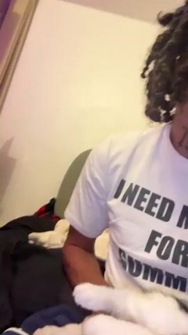 The video shows a person sitting on a bed, with a cat lying on their lap. The person is wearing a white t-shirt with text on it. The cat is black and white and appears to be asleep or resting. The room is dimly lit, and the background is blurry.