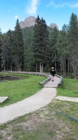 A young person in a black hoodie and shorts walks across a wooden bridge over a path. The bridge is made of rough logs and planks. The path is gravel. A lake is to the left of the bridge, with lily pads on its surface. Tall evergreen trees surround the lake and the bridge, and a mountain peak is visible in the background. The sky is blue with a few white clouds. The ground is covered in green grass.