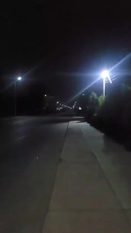 A lone sidewalk runs along a dark road at night. Streetlights illuminate the scene, casting long shadows. The road appears wet, possibly from rain. Trees and bushes line the right side of the road, creating a dark, natural border. The sky is dark and starless, indicating a cloudy or moonless night. The overall impression is one of solitude and quiet.