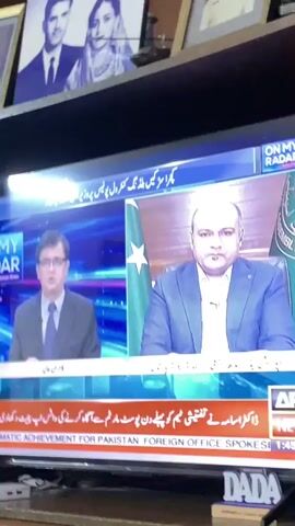 The video shows a television screen displaying a news broadcast. On the left side of the screen is a man wearing a suit and tie, speaking. On the right side of the screen is another man, also in a suit, who appears to be a guest or interviewee. A Pakistani flag is visible behind him. A news ticker at the bottom of the screen displays text in English, including "BREAKING NEWS" and "ACHIEVEMENT FOR PAKISTAN: FOREIGN OFFICE SPOKESPERSON". The overall setting appears to be a news studio.