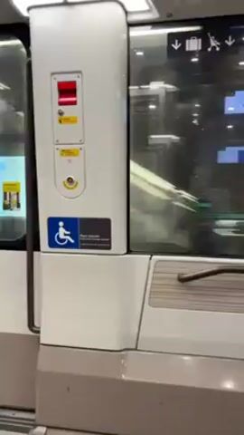 The video shows the interior of a train car, focused on a set of sliding doors. The doors are white and gray with a large window panel. A red light is visible above a yellow button, and a wheelchair accessibility symbol is on the lower part of the door. The train appears to be moving, as the view through the window shows a blur of passing structures.