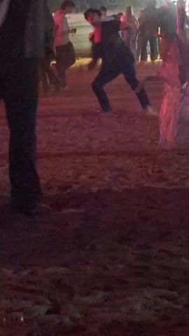 People are walking around a dirt area. The lighting is dim, and there's red and blue light present. Some people are standing still in the background. One person is running in the foreground.