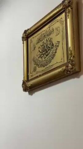 The camera pans up the wall in an interior space revealing three gold-framed pictures hanging on the wall, including one with Arabic calligraphy. Below the pictures, a row of ornate gold chairs or seating is partially visible. The ceiling is white with a decorative cutout section near the upper left.