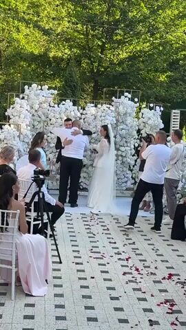The video shows an outdoor wedding ceremony. A bride and groom are standing in front of a backdrop of white flowers. A man in a white shirt and black pants, possibly the groom's father, is hugging the groom. A woman in a light blue dress is holding a basket and walking. A woman is speaking into a microphone. Several people are filming the ceremony with cameras and phones. Guests are seated in chairs on the right side of the frame. The ground is paved with square tiles and scattered with rose petals. Green trees are visible in the background.