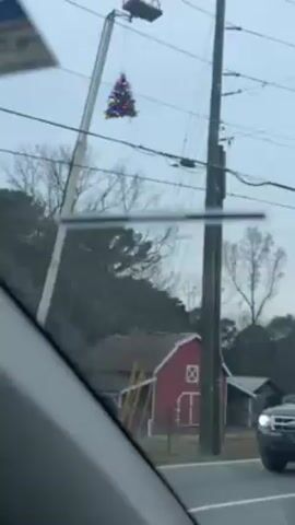 The video shows a decorated Christmas tree on top of a utility pole or very tall pole along a road. The video is taken from inside a moving vehicle. In the background is a red barn and trees. The sky is overcast.