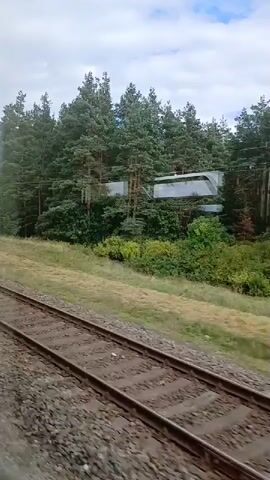This video shows a view from a moving train. The train tracks are in the foreground, and the landscape to the right is blurred due to motion. The landscape consists of a grassy area, a line of trees, and some modern buildings or structures partially obscured by the trees. The sky is cloudy.