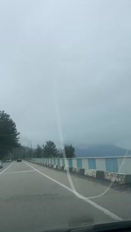 The video shows a car driving on a road with a bridge. The weather is cloudy and the sky is overcast. There are trees on the left side of the road. The bridge has a blue and white railing. There is a car ahead on the road. There are some mountains in the background covered in fog. The asphalt of the road is gray.