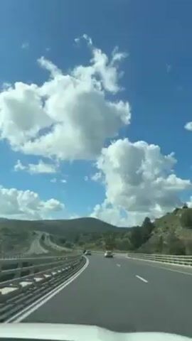 The video shows a view from a car driving on a highway, with rolling green hills in the background and a bright blue sky filled with fluffy white clouds overhead. There are several cars visible on the highway, moving in the same direction as the camera car. Streetlights line the side of the road. A green sign with directions is briefly visible. The road curves gently, following the terrain. The overall impression is a sunny day on a scenic drive.