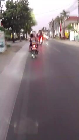 The video captures a moment from the perspective of someone riding on the back of a motorcycle. The rider is looking ahead down a paved road lined with trees and buildings. Several other motorcycles and a car are visible, all moving in the same direction. The sky appears to be dusky, suggesting it is either dawn or dusk. The scene is bustling with activity, typical of a street in a populated area. The focus is on the movement of the vehicles and the general atmosphere of the roadway.