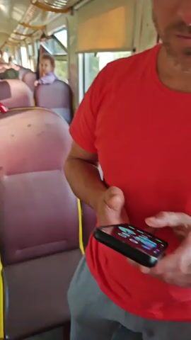 The video shows the interior of a train car. The seats are a deep burgundy with yellow trim. Several passengers are visible. In the foreground, a person wearing a red shirt is partially visible. Further down the aisle, a young child wearing a white shirt and blue pants walks away from the camera. A young girl with blonde hair sits in a seat near the window, looking towards the camera. An adult woman with blonde hair and sunglasses sits by the window, looking out. The lighting is bright, suggesting daylight.