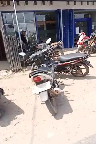 The video opens with a shot of a dusty street in front of several buildings. Several motorcycles are parked in front of one of the buildings, which appears to be a shop with a blue facade and large glass doors. To the right, a line of motorcycles is parked, some of which are being loaded into a vehicle. Further down the street, a wheelbarrow filled with cement is visible, suggesting construction or renovation work is underway. The video then pans across the street, showing more buildings and a person sitting near an open doorway. A black motorcycle is parked in the foreground of the scene. The overall environment appears to be a developing urban or semi-urban area.