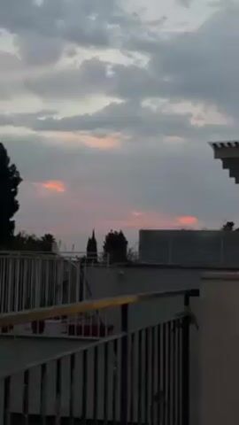 The video shows a view from a building's rooftop, featuring a cloudy sky at sunset with patches of red light breaking through. The foreground includes a white railing surrounding the rooftop area. Beyond the railing, there are some plants in pots and taller trees in the distance. The sky is predominantly gray with the reddish hues of the setting sun creating a contrast near the horizon. The scene appears tranquil, capturing a moment of dusk.