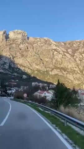 The camera pans slowly from left to right while on a vehicle traveling along a paved road. The road has a metal guard rail. In the distance is a large, rocky mountain under a clear, blue sky. The mountain is illuminated by sunlight, creating shadows on its surface. At the foot of the mountain are several houses with red roofs. As the camera pans, bushes and trees appear in the foreground, partially obscuring the mountain view.